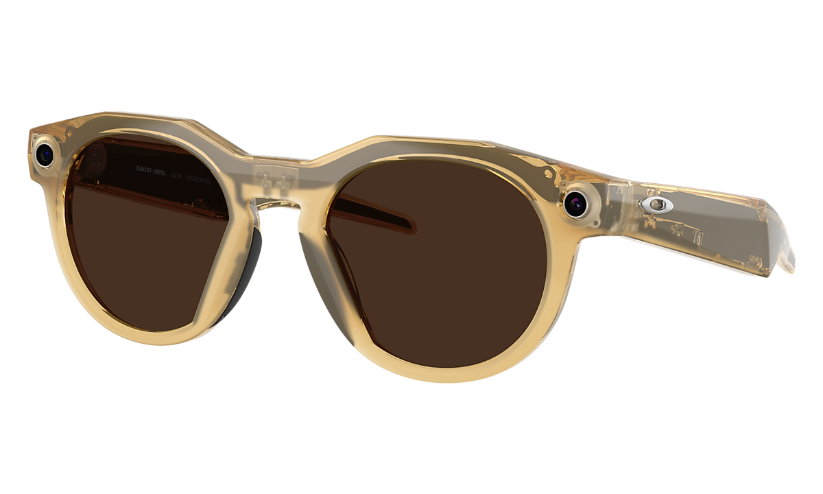 Oakley Meta HSTN AI glasses in a translucent amber frame with brown lenses, showing the built-in camera lens
